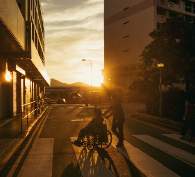 A person pushing a wheelchair along a quiet street at sunset in Singapore, capturing the quiet weight of caregiving.