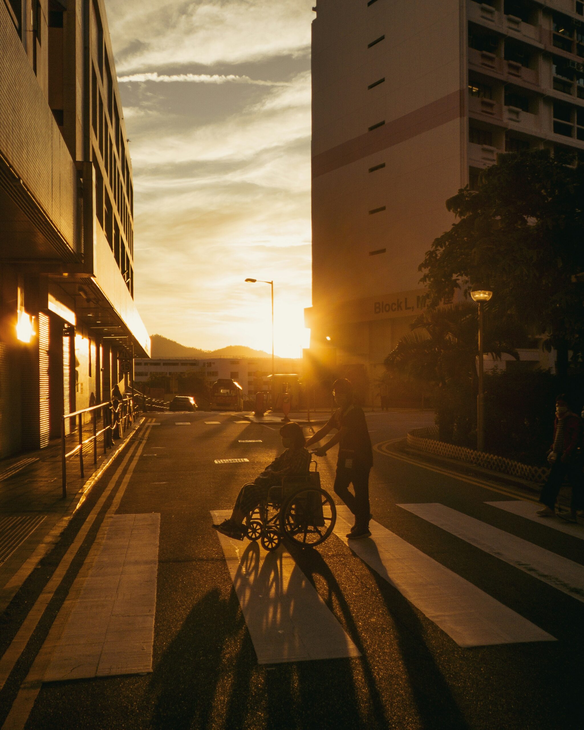 A person pushing a wheelchair along a quiet street at sunset in Singapore, capturing the quiet weight of caregiving.