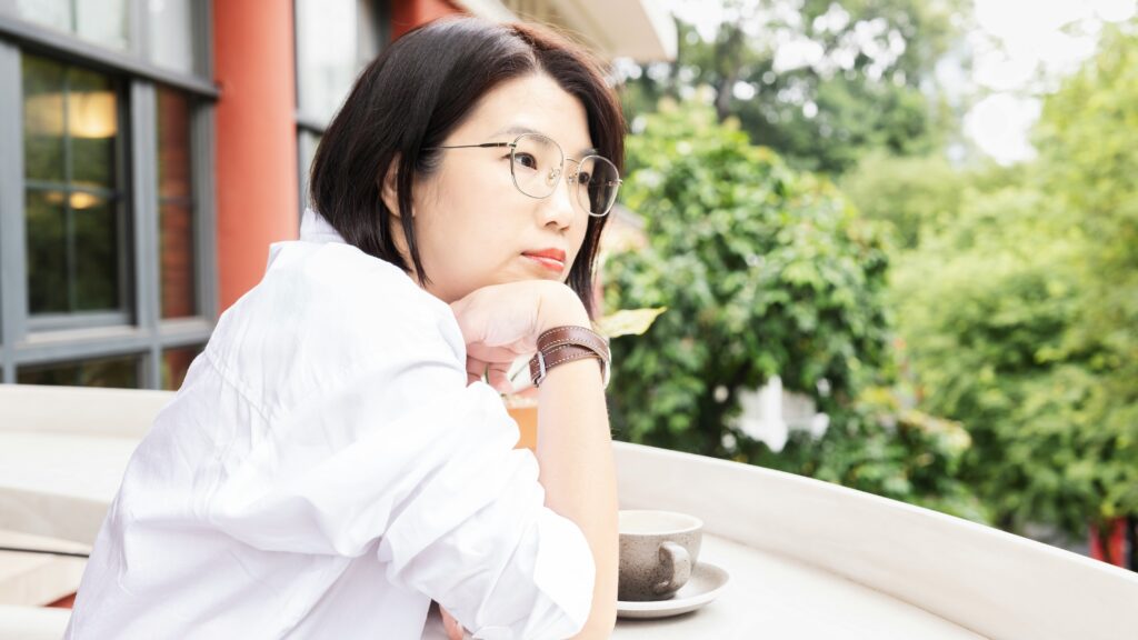 A woman in a white shirt sits at an outdoor table gazing thoughtfully into the distance, capturing the quiet inner weight of never feeling good enough.