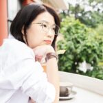 A woman in a white shirt sits at an outdoor table gazing thoughtfully into the distance, capturing the quiet inner weight of never feeling good enough.