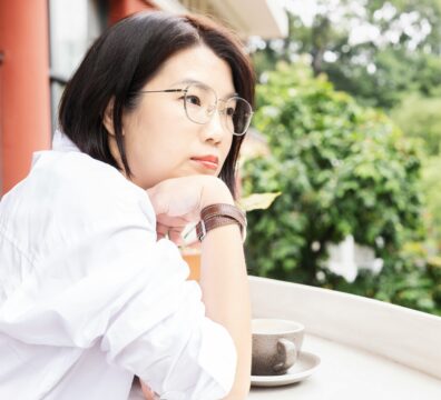 A woman in a white shirt sits at an outdoor table gazing thoughtfully into the distance, capturing the quiet inner weight of never feeling good enough.