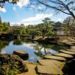 Stepping stones cross a still pond in a Japanese garden, evoking the quiet, deliberate process of grounding and finding your way back to calm.