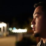 A man alone at night gazes quietly into the distance, his face softly lit against a backdrop of blurred city lights, capturing the experience of carrying something privately while appearing composed to the world.