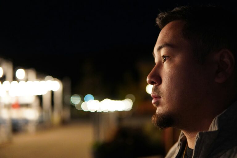 A man alone at night gazes quietly into the distance, his face softly lit against a backdrop of blurred city lights, capturing the experience of carrying something privately while appearing composed to the world.
