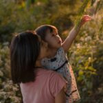 A mother holds a young child in warm golden light as the child reaches upward toward green branches, evoking the safety and support of being held while reaching toward something new — the essence of reparenting.