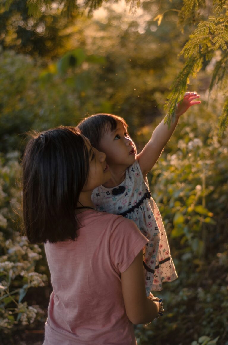 A mother holds a young child in warm golden light as the child reaches upward toward green branches, evoking the safety and support of being held while reaching toward something new — the essence of reparenting.