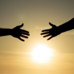 Two hands reach toward each other in silhouette against a warm golden sunset, with a gap between them that evokes the longing for connection and the distance that attachment patterns can create.