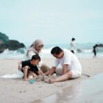 A family of three — mother, father, and young child — play together in the sand at a beach, capturing the warm and visible moments of parenting that sit alongside the invisible mental load the article explores.