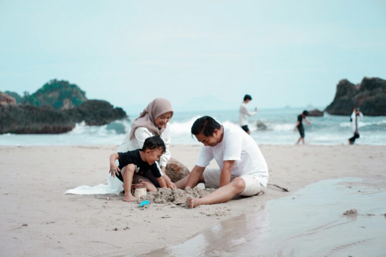 A family of three — mother, father, and young child — play together in the sand at a beach, capturing the warm and visible moments of parenting that sit alongside the invisible mental load the article explores.