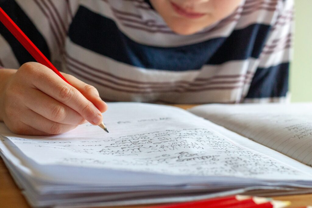 A young child writes in a notebook with a red pencil, focused on their schoolwork, capturing the ordinary but pressured experience of study season for Singapore families.