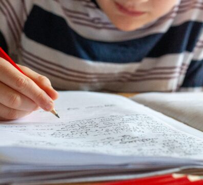 A young child writes in a notebook with a red pencil, focused on their schoolwork, capturing the ordinary but pressured experience of study season for Singapore families.