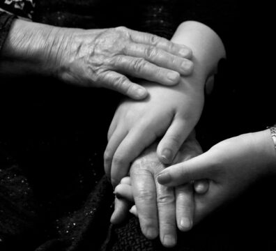 Different generations of hands rest together, an elderly person's hands over a younger adult's hands, quietly capturing the experience of the sandwich generation holding both the older and younger generation at once.