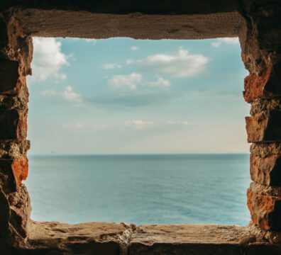 A square stone window in a dark wall opening onto a calm blue sea and sky.