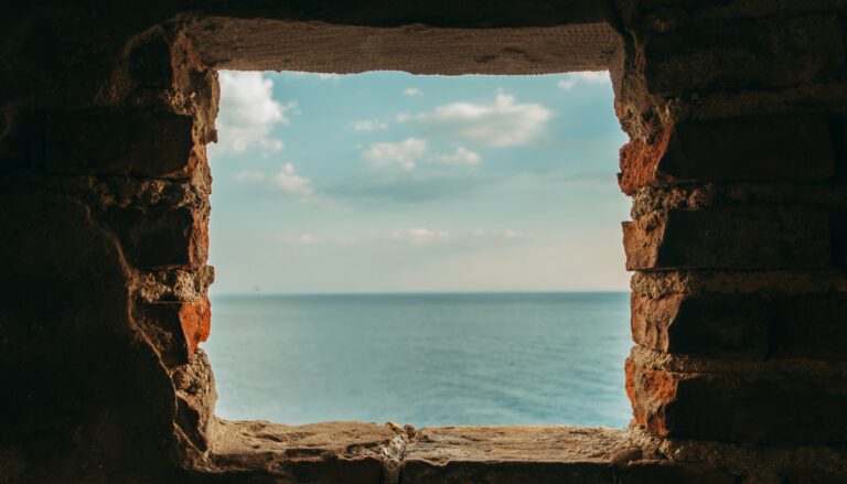 A square stone window in a dark wall opening onto a calm blue sea and sky.