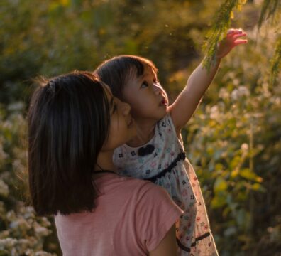 A mother holds a young child in warm golden light as the child reaches upward toward green branches, evoking the safety and support of being held while reaching toward something new — the essence of reparenting.