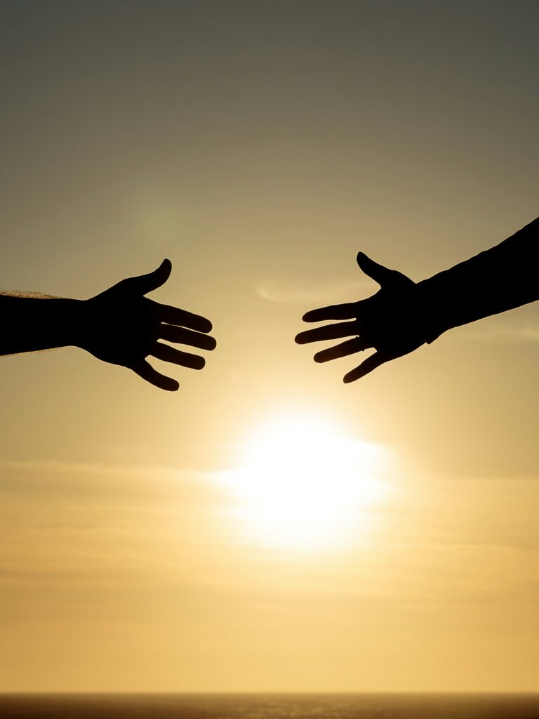 Two hands reach toward each other in silhouette against a warm golden sunset, with a gap between them that evokes the longing for connection and the distance that attachment patterns can create.