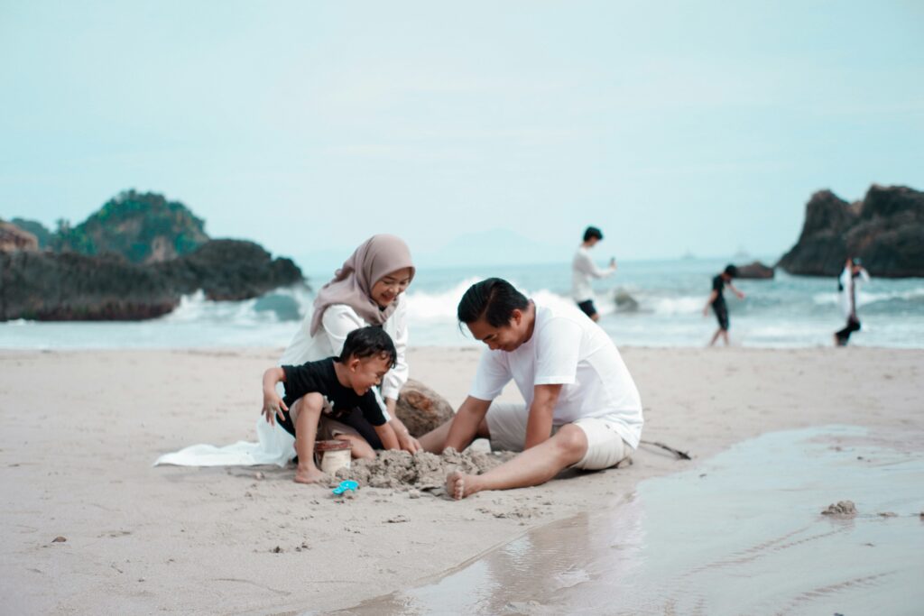A family of three — mother, father, and young child — play together in the sand at a beach, capturing the warm and visible moments of parenting that sit alongside the invisible mental load the article explores.