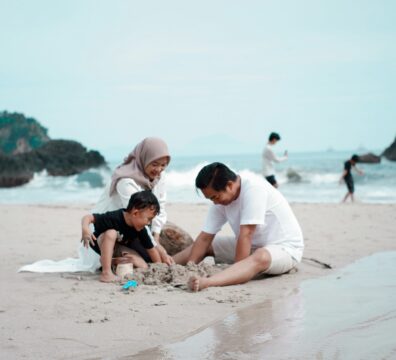 A family of three — mother, father, and young child — play together in the sand at a beach, capturing the warm and visible moments of parenting that sit alongside the invisible mental load the article explores.