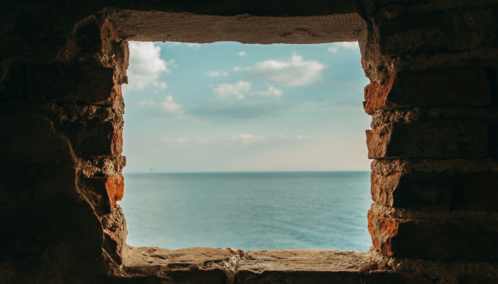 A square stone window in a dark wall opening onto a calm blue sea and sky.